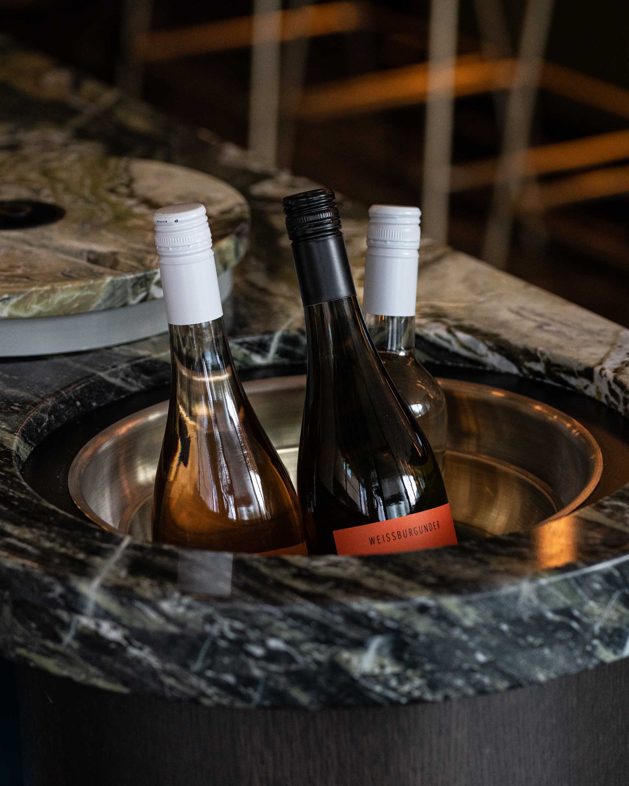 Three bottles of wine chill in a metal bucket within a dark stone bar top