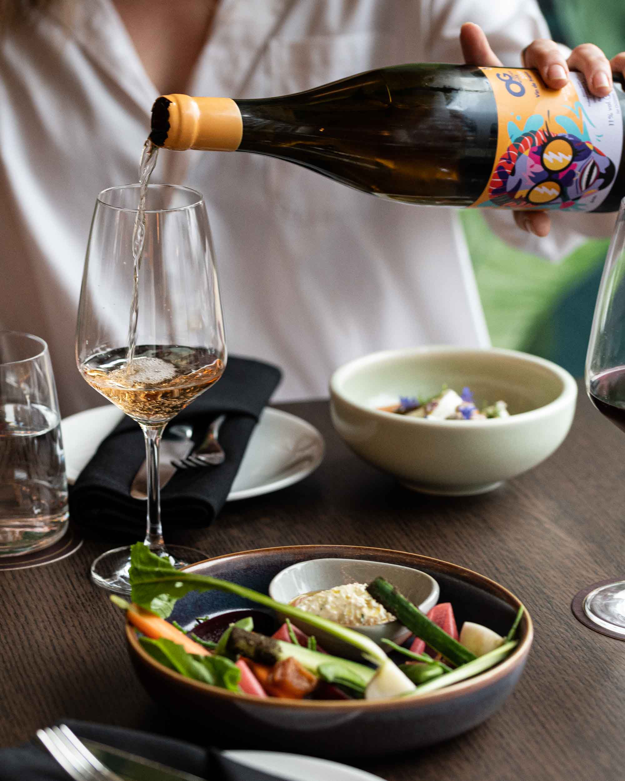 Woman at a table pours a glass of white wine behind fermented, fresh and cured local vegetables in a dark blue bowl