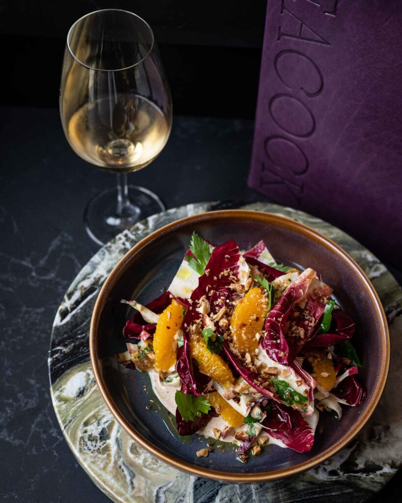 Treviso, ajo blanco, citrus and almond crumble in a dark grey bowl in front of a purple menu and wine glass