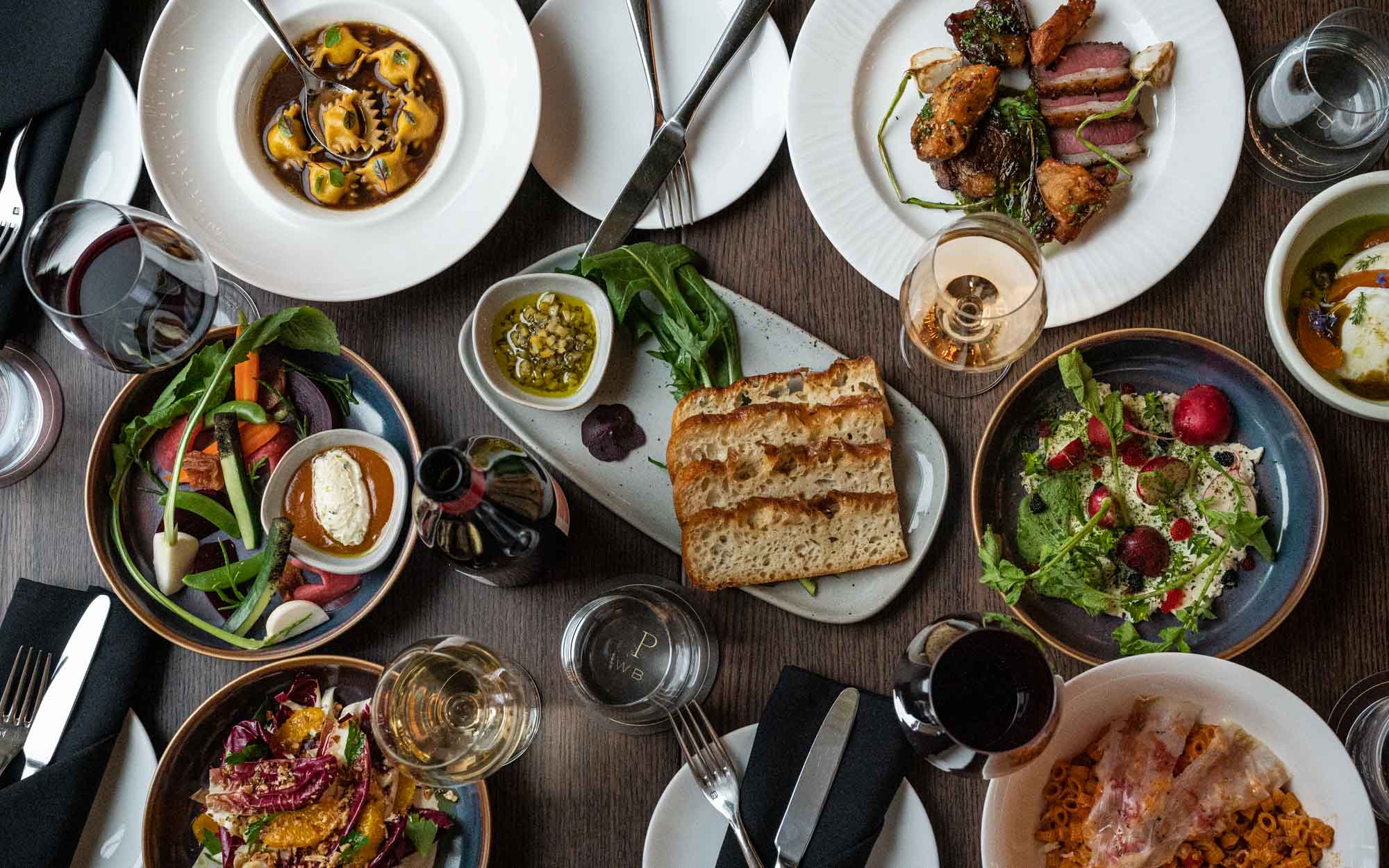 Top view of a table set with wine glasses, focaccia, bavette steak, caviar butter, buffalo mozzarella and fermented, fresh and cured local vegetables